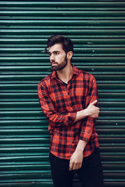 Homem barbudo jovem, modelo de moda, vestindo uma camisa xadrez com um verde cego atrás dele . — Fotografia de Stock