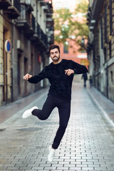 Junger bärtiger Mann springt mit offenen Armen im städtischen Hintergrund und trägt lässige Kleidung. — Stockfoto