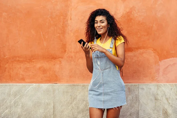 Mujer árabe feliz escuchando música con auriculares contra la pared roja —  Fotos de Stock