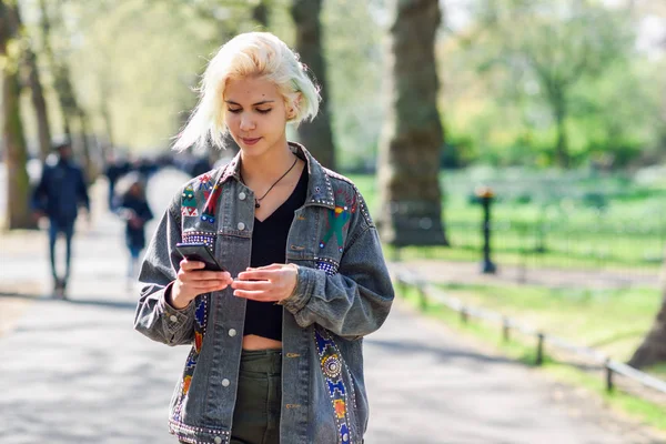 Jeune femme urbaine à la coiffure moderne utilisant un smartphone marchant dans la rue dans un parc urbain . — Photo