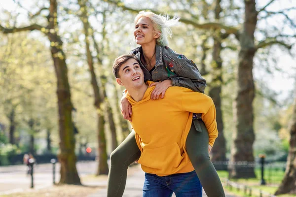 Grappig paar in een stedelijk park. Vriendje het dragen van zijn vriendin op piggyback. — Stockfoto