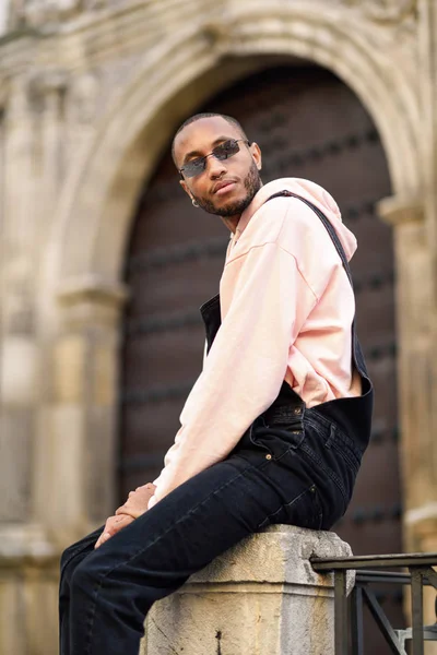 Joven hombre negro con ropa casual y gafas de sol sentado en la calle —  Fotos de Stock
