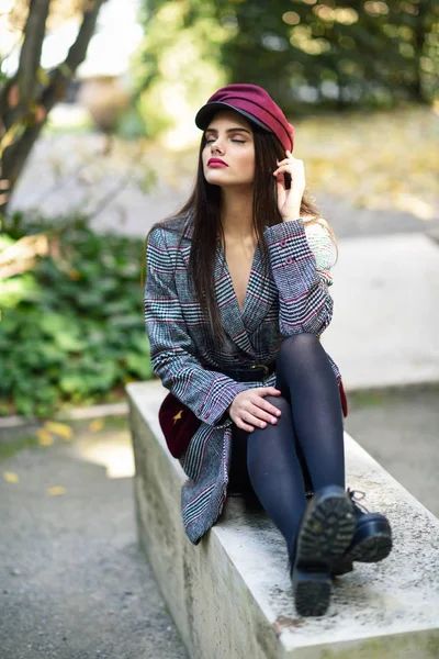 Joven hermosa chica con abrigo de invierno y gorra sentado en un banco en el parque urbano —  Fotos de Stock