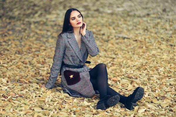 Chica joven con el pelo muy largo con abrigo de invierno sentado en el suelo lleno de hojas de otoño —  Fotos de Stock