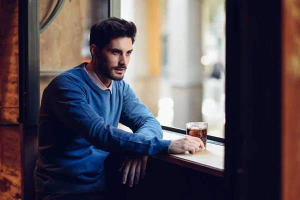 Pensive guy with modern hairstyle near a window drinking a soda. — Stock Photo, Image