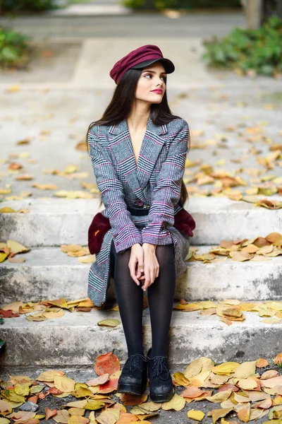 Joven hermosa chica con abrigo de invierno y gorra sentada en escalones llenos de hojas de otoño —  Fotos de Stock