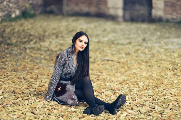 Hermosa chica con abrigo de invierno sentado en el suelo de un parque urbano lleno de hojas de otoño . — Foto de Stock