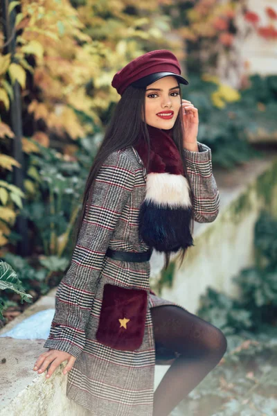 Chica joven con el pelo muy largo sonriendo y usando abrigo de invierno y gorra en el fondo de las hojas de otoño . — Foto de Stock