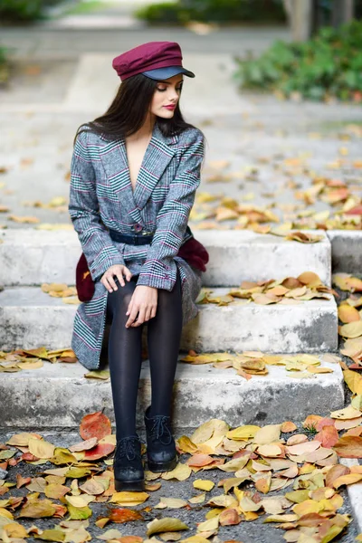 Junge schöne Mädchen in Wintermantel und Mütze sitzen auf Stufen voller Herbstblätter — Stockfoto