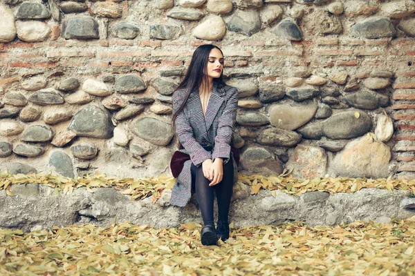 Hermosa chica con abrigo de invierno sentado en el suelo de un parque urbano lleno de hojas de otoño . — Foto de Stock