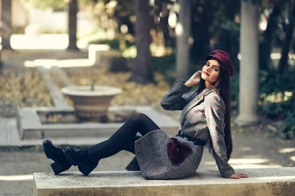 Joven hermosa chica con abrigo de invierno y gorra sentado en un banco en el parque urbano . — Foto de Stock