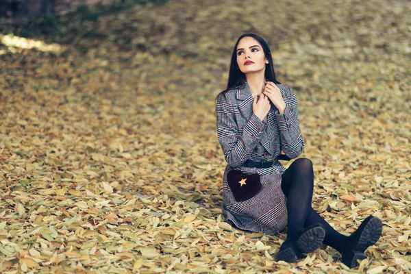 Vacker flicka klädd i vinterpäls sitter på golvet i en Urban Park full av höstlöv. — Stockfoto
