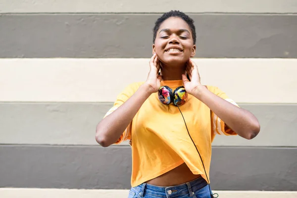 Mujer africana feliz sonriendo en la pared urbana con los ojos cerrados, con ropa casual y auriculares . —  Fotos de Stock