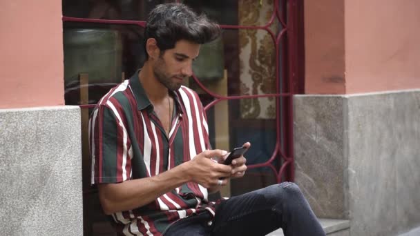 Jeune homme utilisant un téléphone intelligent assis sur l'étape urbaine . — Video