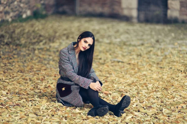 Piękna dziewczyna ubrana w płaszcz zimowy siedzi na podłodze parku miejskiego pełnego jesiennych liści. — Zdjęcie stockowe
