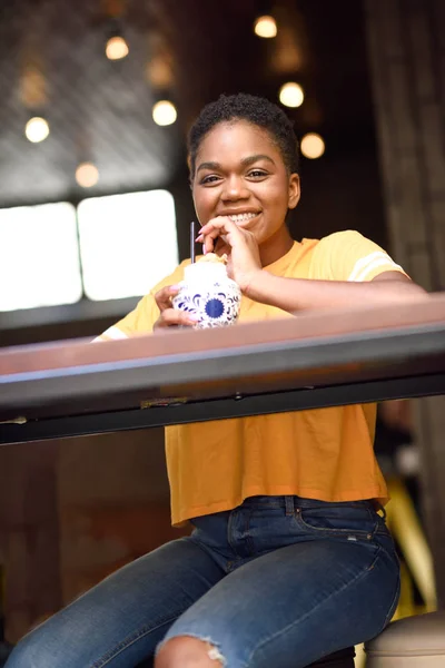 Sonriente chica negra con el pelo muy corto bebiendo un cóctel en un café urbano . — Foto de Stock