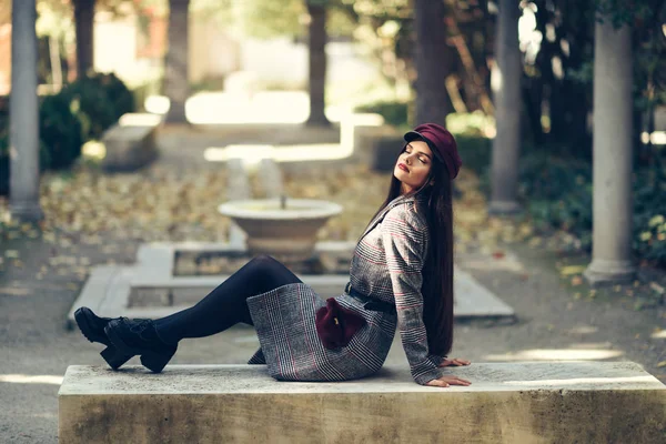 年轻的漂亮女孩穿着冬衣和帽子坐在城市公园的长椅上. — 图库照片