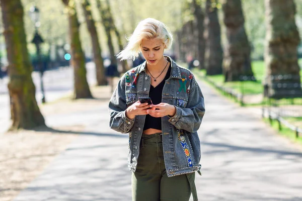 Modern saç modeli Ile genç kentsel kadın bir kentsel park sokakta yürüyüş Smartphone kullanarak. — Stok fotoğraf