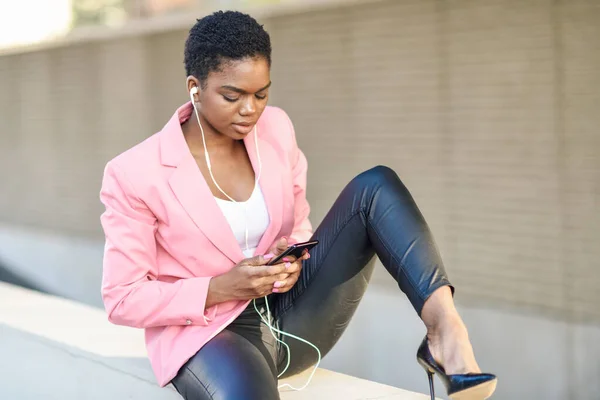 Empresaria negra sentada al aire libre usando smartphone con auriculares —  Fotos de Stock