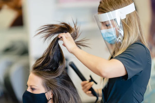 Friseurin, geschützt durch eine Maske, kämmt in einem Salon die Haare ihrer Kunden mit einem Haarbügeleisen. — Stockfoto
