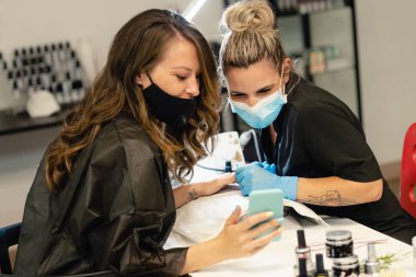 Müşteri akıllı telefonuyla güzellik uzmanına fotoğraf gösteriyor. İkisi de maskelerle korunuyor..