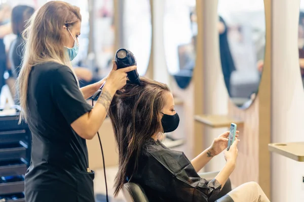 Cabeleireiro secando seus clientes cabelo com um secador de cabelo usando máscaras protetoras em um centro de beleza. — Fotografia de Stock