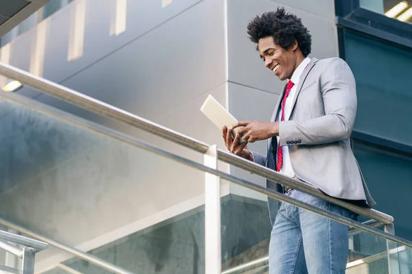 Hombre de negocios negro usando una tableta digital sentada cerca de un edificio de oficinas. —  Fotos de Stock