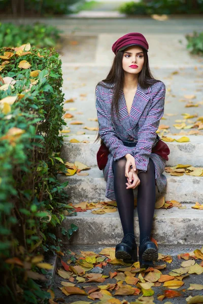 Joven hermosa chica con abrigo de invierno y gorra sentada en escalones llenos de hojas de otoño — Foto de Stock