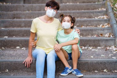 Orta yaşlı anne ve kız sokakta maske takarak oturuyorlar.