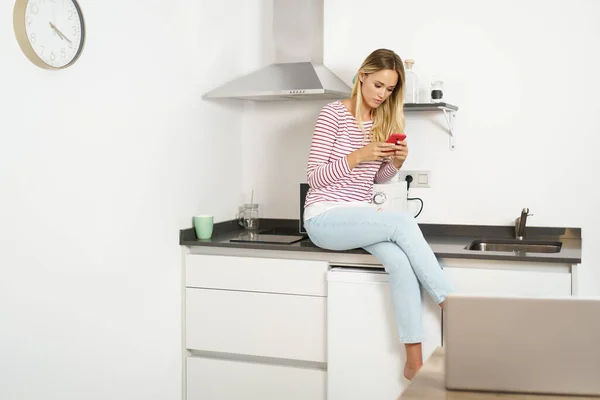 Pensando hermosa mujer usando su teléfono inteligente sentado en la cocina en casa —  Fotos de Stock