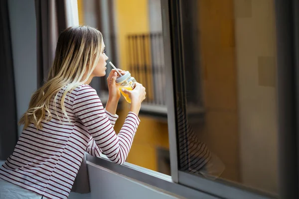 Mladá žena pije sklenici přírodní pomerančové šťávy, naklání se z okna svého domu. — Stock fotografie
