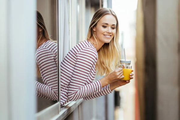 Lächelnde Frau trinkt ein Glas natürlichen Orangensaft und lehnt sich aus dem Fenster ihres Hauses. — Stockfoto