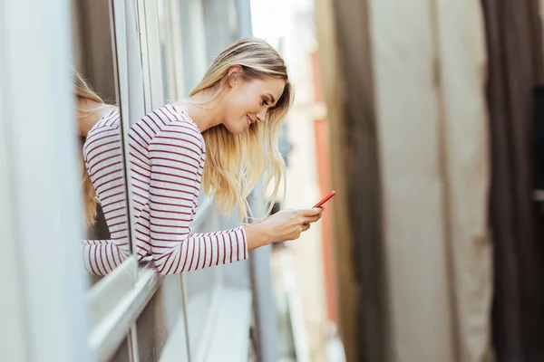 Blanke vrouw leunend uit haar huis raam met behulp van een smartphone. — Stockfoto