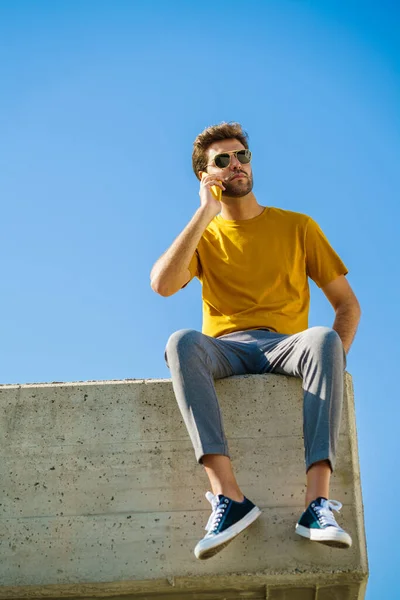 Młody człowiek za pomocą smartfona siedzi na parapecie na zewnątrz — Zdjęcie stockowe