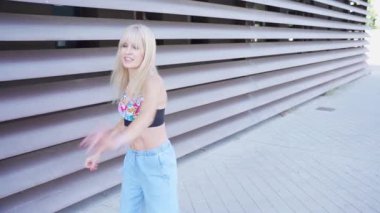 Genç bir kadın modern bir binanın dışında dans etmekten hoşlanıyor. Üzerinde çiçekli desenler, açık renk kot pantolonlar ve kaygısız ve neşeli bir hava olan siyah bir bluz giyiyor..