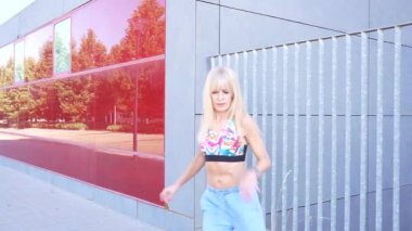 Aktif sarışın kadın şehir ortamında renkli spor sutyen ve kot şort giyerek koşuyor, sağlıklı bir yaşam tarzını kucaklıyor. Büyük pencereli binalar modern bir şehir arkaplanı sağlıyor.