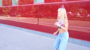 Güneşli bir günde enerjik genç bir kadın enerjik bir şekilde kırmızı bir duvarın önünde dans eder, neşe ve özgürlüğü ifade eder. Ekin bir bluz ve kot pantolon giyiyor, modern ve modaya uygun bir tarzı sergiliyor.