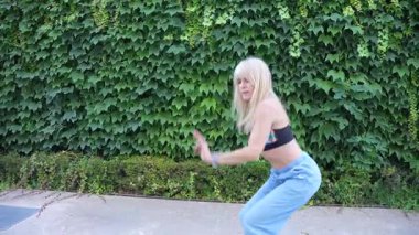 Sarışın bir kadın açık havada enerjik bir şekilde dans ediyor. Kırpılmış bluz ve kot pantolon giyiyor. Neşeli hareketleri enerjik ve dinamik bir havayı yansıtıyor. Eğlenceli ve özgür bir tavır sergiliyor.