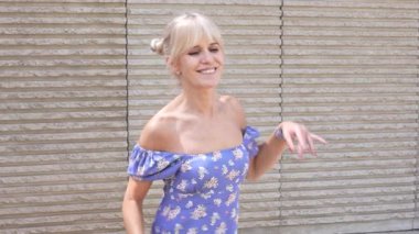 Şık bir çiçekli yaz elbisesi içinde çekici bir sarışın kadın gün ışığında şehir zeminine karşı mutlu bir şekilde poz verir. Kaygısız tavırları ve parlak ifadesiyle neşe ve neşe saçar.