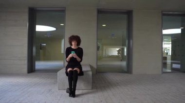 Kıvırcık saçlı, şık siyah elbiseli bir kadın, taştan bir bankta rahat bir şekilde oturuyor, akıllı telefonuyla derinden meşgulken, şık, minimalist mimari bir arka planla çevrili.