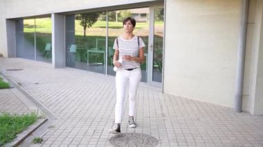 Bir kadın, şık çizgili bir bluz ve çıtır çıtır beyaz bir pantolon giymiş, modern mimarinin çağdaş güzelliği ile modayı kusursuz bir şekilde birleştiren, güvenli bir şehir manzarasında adımlar atar.