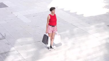 Şık bir birey, canlı bir şehir ortamında birkaç alışveriş torbası tutarken, parlak güneş ışığı altında canlı ve renkli yaz kıyafetlerini gururla sergilerken, güven içinde ayakta duruyor.