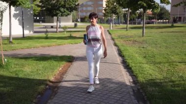 Şık bir atletizm elbisesi giymiş şık bir genç kadın, güneşli ve parlak bir parkta rahatça geziniyor, modern moda ve aktif, fitnesiyeli bir yaşam tarzı karışımı sergiliyor.