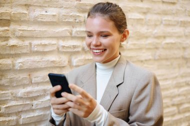 Şık bir spor ceketli neşeli genç bir kadın, akıllı telefonuyla dokulu bir duvara karşı etkileşime giriyor, bir anlık mutluluk ve dijital dünyayla bağlantı kuruyor, modaya uygun bir yaşam tarzını somutlaştırıyor.