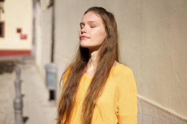 Genç bir kadın güneşli bir sokakta dikiliyor, gözleri kapalı, güneşin sıcacık ve ışıl ışıl parlayan parıltısıyla güneşlenirken, aynı zamanda neşeli ve neşeli sarı bir kazak giyiyor ve bu ona neşe katıyor.