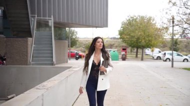 Kendine güvenen genç bir kadın canlı bir şehir ortamında yürüyor. Şık ve şık giyiniyor. Çevresi yeşillik ve modern binalarla çevrili.