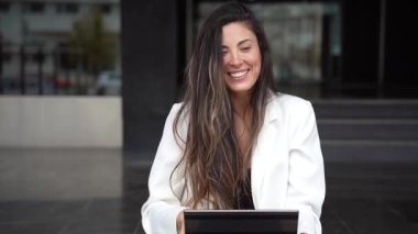 Süslü beyaz bir ceket giymiş genç bir kadın dışarıda eğleniyor. Modern bir tablet kullanırken rahat bir şekilde oturuyor. Gelişigüzel bir yaşam tarzıyla teknolojinin çekici bir karışımını sergiliyor.