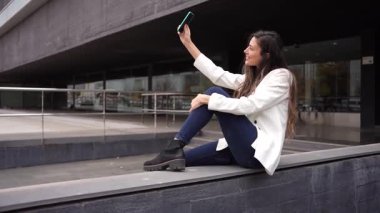 Tarz sahibi genç bir kadın pervazda poz veriyor, akıllı telefonuyla mükemmel bir selfie çekiyor. Çevresindeki modern mimari, canlı şehir atmosferini geliştiriyor ve onu ideal bir zemin haline getiriyor.