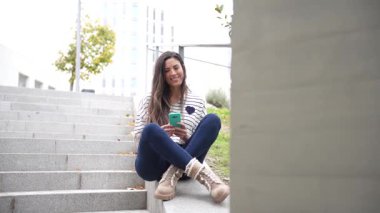 Genç bir kadın, kentteki modern bir şehir yaşam tarzını güzel bir şekilde sergileyen akıllı telefonuyla, açık hava basamaklarında otururken bir anlık dinlenmenin tadını çıkarıyor.