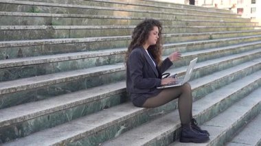 Genç bir kadın yıpranmış taş basamaklara oturur, dizüstü bilgisayarına odaklanır, modern bir çalışma alanını canlı bir dış mekan ortamı ile harmanlayarak, hem doğaya hem de teknolojiye olan bağlılığını gösterir.